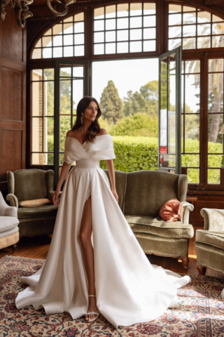 Brautkleid Bryony von vorne: Elegante, cleane A-Linie aus Satin mit markantem breiten Bund an der Taille. Der Rock fällt fließend und verfügt über einen hohen Beinschlitz. Die voluminösen Off-Shoulder-Ärmel setzen ein modernes Statement