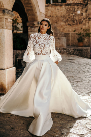 Brautkleid Rhea: Zweiteiliges Hochzeitskleid mit Satinrock in Ivory, tiefen Kellerfalten und breitem Bund. Transparentes Oberteil aus perlenbestickter Spitze mit angedeutetem Rollkragen und weit ausgestellten Ärmeln.